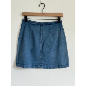 May include: A light blue denim skirt hanging on a wooden hanger. The skirt is a classic A-line style with a button and zipper closure. The skirt is made of a medium-wash denim fabric.