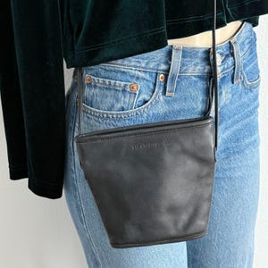 Könnte beinhalten: Eine kleine Umhängetasche aus dunkelbraunem Leder mit einer strukturierten, konisch zulaufenden Eimerform. Auf der Vorderseite der Tasche befindet sich der geprägte Schriftzug Liz Claiborne. Sie hat einen dünnen, passenden Lederriemen.