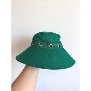 Op de afbeelding: Een groene zonnehoed met een brede rand en een reeks decoratieve metalen oogjes rond de kroon. De hoed is gemaakt van effen groene stof en heeft een gestikt ontwerp op de rand.