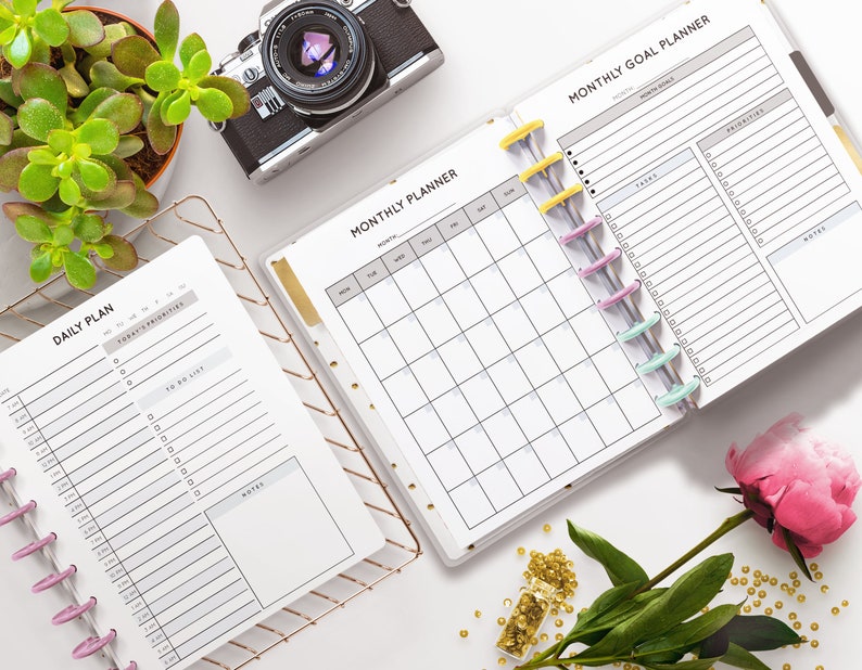 May include: A white planner with a monthly calendar and a daily planner. The planner has a gold ring binder and is open to the monthly calendar page. The daily planner is open to the daily page. The planner is on a white surface with a succulent plant, a camera, a flower, and gold beads.