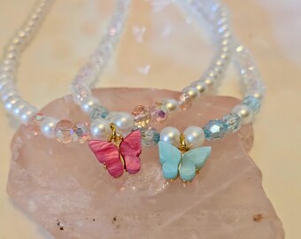 May include: Two delicate necklaces with white pearl beads and butterfly pendants. One butterfly is pink, the other is light blue. The necklaces rest on a pink, textured surface. The butterfly pendants are attached with gold-colored metal.