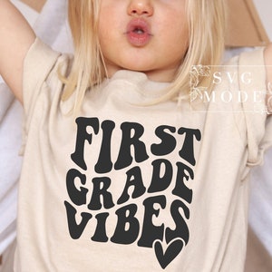 May include: A beige t-shirt with the words "FIRST GRADE VIBES" in a black, retro font. A small black heart is below the word "VIBES". The shirt is a short-sleeved crew neck.