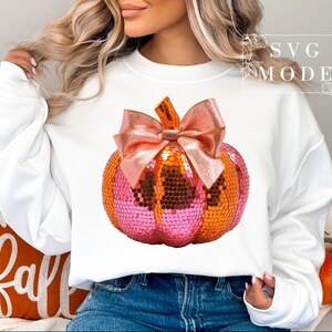 Peut inclure: Un sweat-shirt blanc avec une citrouille en sequins rose et orange surmontée d'un nœud rose. Le sweat-shirt a un col rond et des manches longues.