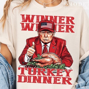 May include: A white t-shirt with a red, white, and blue graphic of a man wearing a red suit and a red, white, and blue baseball cap. The graphic also includes a turkey and the text "Winner Winner Turkey Dinner".