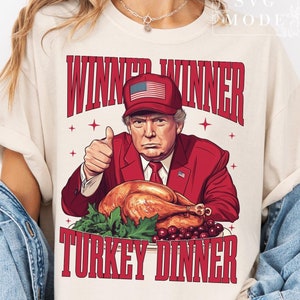 May include: Cream-colored t-shirt with a graphic of a man in a red suit and a hat with an American flag, giving a thumbs-up. The text "WINNER WINNER TURKEY DINNER" is printed in red.