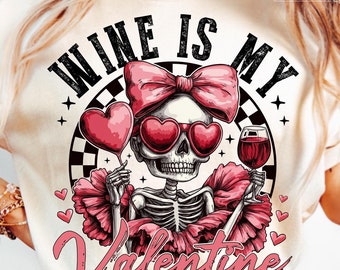 May include: A white t-shirt with the text "Wine is my Valentine" in black lettering. A pink bow and skull with sunglasses and a glass of wine are featured in the design.