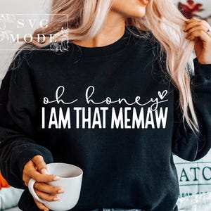 May include: Black sweatshirt with the text "oh honey I AM THAT MEMAW" in white script. The sweatshirt is a crew neck style. A white coffee mug is held in the hand. The image is of a person.