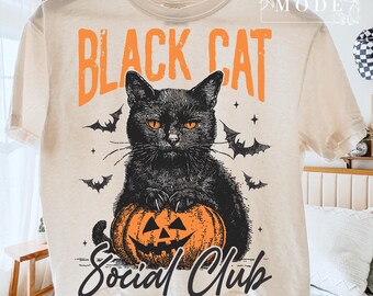 May include: Cream-colored t-shirt featuring a black cat with orange eyes, holding an orange jack-o'-lantern. The text "BLACK CAT" is above the cat in orange, and "Social Club" is below in black script. Bats and stars are also featured.