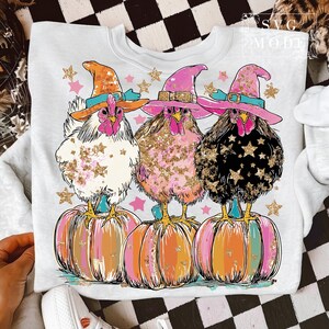 May include: A white sweatshirt featuring three chickens wearing witch hats and standing on pumpkins. The chickens are decorated with gold glitter and stars. The pumpkins are painted in shades of orange, pink, and yellow. Pink and gold stars are scattered around the chickens.
