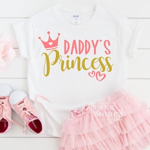 May include: White t-shirt with a pink crown and gold lettering that says "Daddy's Princess" with two pink hearts. The shirt is paired with a pink tulle skirt and pink sneakers.