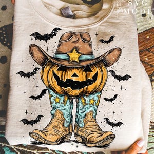May include: Beige sweatshirt featuring a Halloween-themed design. The graphic includes a carved pumpkin wearing a cowboy hat with a star, cowboy boots, and bats. The design is in shades of orange, brown, and blue.