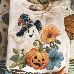 May include: A beige sweatshirt featuring a Halloween-themed design. The design includes a smiling ghost wearing a floral hat, a jack-o'-lantern, and colorful flowers. The sweatshirt is folded, with the design prominently displayed.