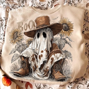 May include: Beige sweatshirt featuring a ghost wearing a cowboy hat and boots, with the word "Boo" above. The ghost is surrounded by sunflowers. The design has a vintage, hand-drawn style, perfect for Halloween.