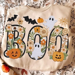 May include: Beige sweatshirt with the word "BOO" embroidered in black, decorated with flowers, pumpkins, bats, ghosts, and a spiderweb. The letters are filled with floral designs and pumpkins. The overall theme is Halloween.