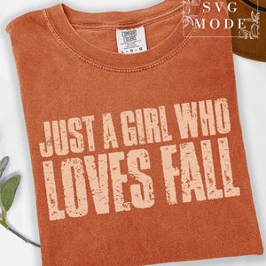 May include: Burnt orange t-shirt with the text "JUST A GIRL WHO LOVES FALL" in a distressed, light orange font. The shirt is folded and has a Comfort Colors tag. A brown hat is visible in the background.
