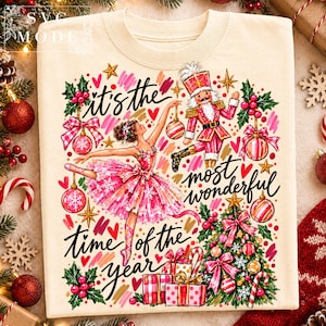 May include: A cream-coloured crewneck sweatshirt featuring a colourful graphic print of a ballerina, a nutcracker, a Christmas tree, and presents. The text reads, It's the most wonderful time of the year, surrounded by holly, ornaments, and stars.