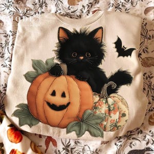 May include: A cream-colored sweatshirt featuring a black cat sitting on a pumpkin with a carved face. A smaller pumpkin with floral patterns and a bat are also depicted. The design is perfect for Halloween.