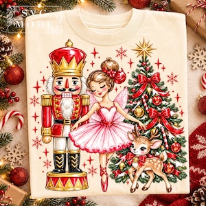May include: A cream-coloured t-shirt featuring a graphic print of a nutcracker, a ballerina in a pink tutu, a decorated Christmas tree, and a fawn. The text "SVG MODE" is printed in the upper left corner of the shirt design.