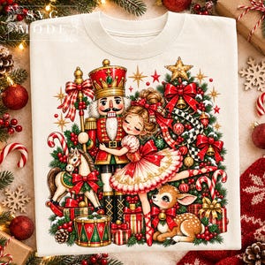 May include: A cream-colored t-shirt featuring a festive graphic of a nutcracker, a ballerina, a rocking horse, and a fawn surrounded by a Christmas tree, presents, and candy canes. The text SVG MODE is printed in the upper left corner of the shirt.