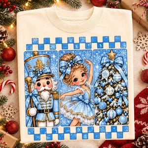 May include: A cream-colored t-shirt featuring a graphic print of a nutcracker, a ballerina, and a decorated Christmas tree in shades of blue. The design is framed by a blue and white checkered border with snowflake accents on a light blue background.
