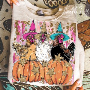 May include: Beige sweatshirt with a design of three chickens in witch hats, standing on orange pumpkins with gold star decorations. The background has pink and gold accents, ideal for Halloween or autumn.