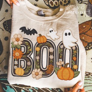 May include: Beige sweatshirt with the word "BOO" in a decorative design featuring a bat, pumpkins, ghosts, flowers, and a spiderweb. The letters are outlined in brown with floral and leaf accents, perfect for Halloween.
