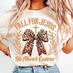 Puede incluir: Una camiseta blanca con el texto "Fall for Jesus" en letras marrones con un lazo de estampado de leopardo y una cruz sobre el lazo. El texto "He Never Leaves" está debajo del lazo.