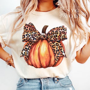 Puede incluir: Una camiseta blanca con un gráfico de una calabaza naranja con un lazo de estampado de leopardo. El texto "SVG MODE" es visible en el fondo.