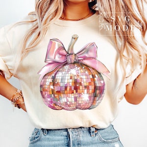 May include: A white t-shirt with a pink and gold glitter pumpkin with a pink bow. The pumpkin is a disco ball design.