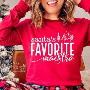 May include: Red long-sleeve shirt with white text that reads "santa's favorite maestra" and a small graphic of Christmas trees.