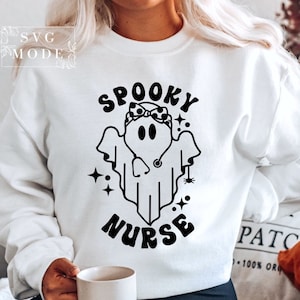 May include: White sweatshirt with a black and white illustration of a ghost wearing a stethoscope and a headband with polka dots. The text "Spooky Nurse" is printed below the ghost.