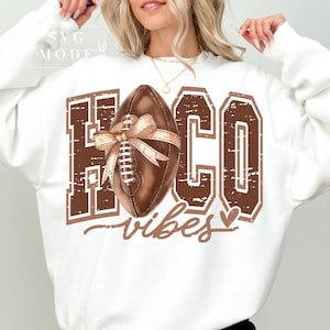 Könnte beinhalten: Weißer Pullover mit einem braunen Football und dem Text "HOCO vibes" in einer verwitterten Schrift.