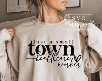 Puede incluir: Una sudadera beige con el texto "Just a small town healthcare worker" en negro con un pequeño gráfico de corazón.