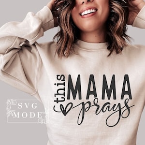 Puede incluir: Una sudadera beige con el texto "this mama prays" en letras negras con un pequeño gráfico de corazón.