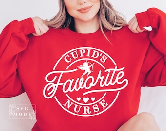 以下が含まれることがあります： 白い円形グラフィックの赤いスウェットシャツ。「Cupid's Favorite Nurse」と書かれており、弓矢を持ったキューピッドと2つのハートが描かれています。スウェットシャツはカジュアルなスタイルです。