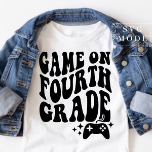 May include: White t-shirt with black text that reads "Game On Fourth Grade" with a video game controller graphic below. The shirt is displayed on a denim jacket.