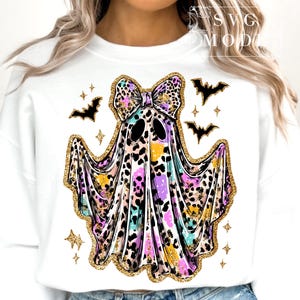 May include: White sweatshirt featuring a colorful ghost design with a bow, leopard print, and gold glitter accents. The ghost has black eyes and is surrounded by black bats and gold stars. A fun Halloween-themed design.