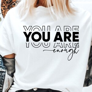 May include: White t-shirt with the text "You are You are You are enough" in black lettering.