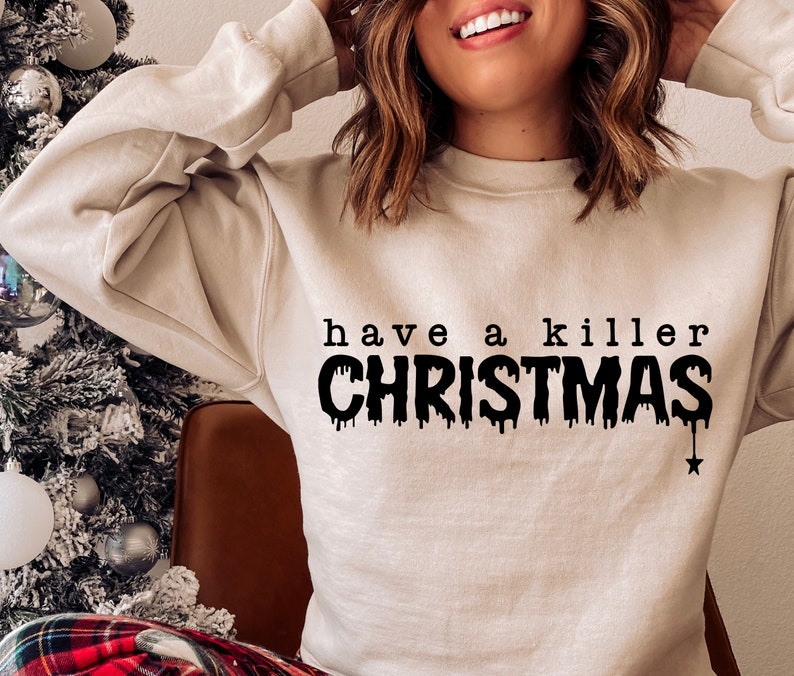 May include: A beige sweatshirt with the phrase "have a killer CHRISTMAS" printed in black. The word "CHRISTMAS" has a dripping effect, and a black star hangs from the bottom of the word. The sweatshirt is a casual holiday garment.