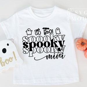 Puede incluir: Camiseta blanca con un dise&ntilde;o gr&aacute;fico negro que dice "Spooky mini" con fantasmas, murci&eacute;lagos y calabazas. El dise&ntilde;o est&aacute; en un estilo superpuesto y en capas.