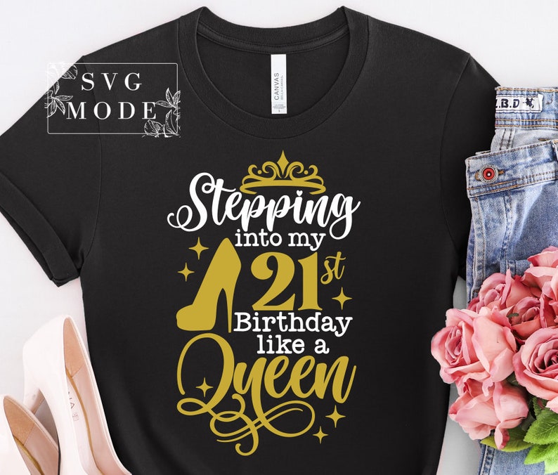 Pode incluir: Camiseta preta com um salto alto dourado e o texto "Stepping into my 21st Birthday like a Queen". O design inclui uma coroa e estrelas. A camisa &eacute; combinada com jeans azuis e rosas cor-de-rosa.