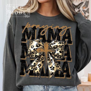 Puede incluir: Sudadera gris oscuro con las palabras "praying MAMA" en un dise&ntilde;o en capas. Un lazo con estampado de leopardo con una cruz en el centro es el punto focal. La sudadera tiene un corte holgado.