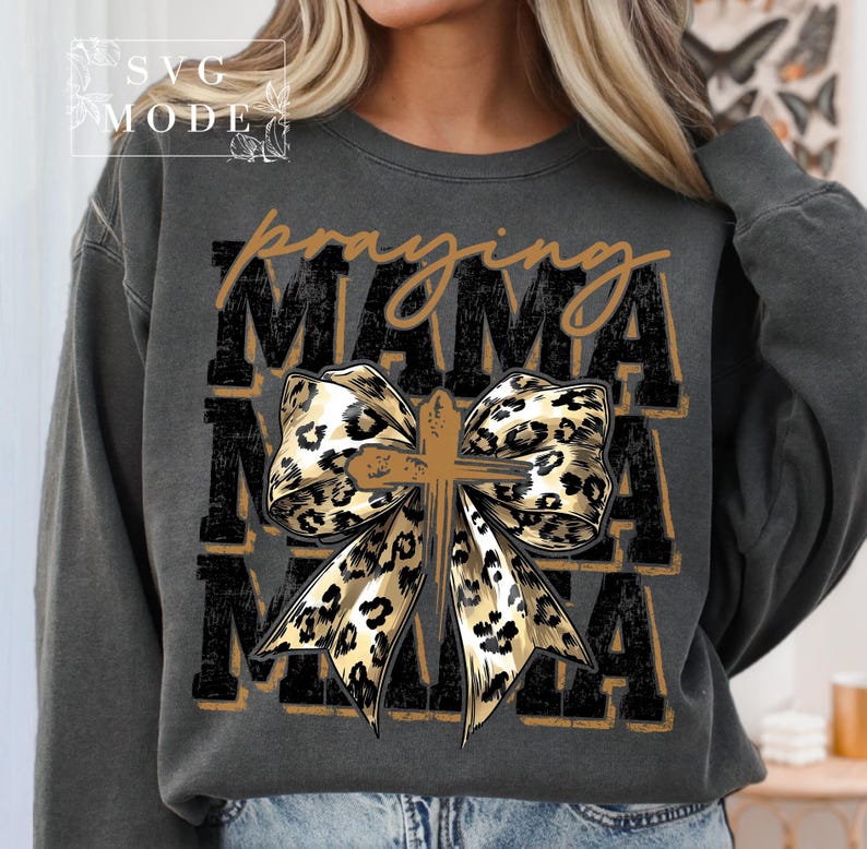 Puede incluir: Sudadera gris oscuro con las palabras "praying MAMA" en un dise&ntilde;o en capas. Un lazo con estampado de leopardo con una cruz en el centro es el punto focal. La sudadera tiene un corte holgado.