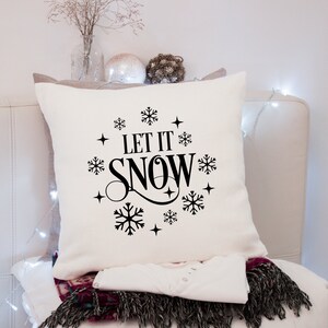 Puede incluir: Un coj&iacute;n cuadrado blanco roto con la frase "LET IT SNOW" en letras negras, rodeada de copos de nieve y estrellas. El coj&iacute;n forma parte de un tema de vacaciones de invierno.