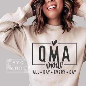 May include: Beige sweatshirt with the text "QMA mode" inside a black rectangle, with a heart above the "Q" and the words "ALL • DAY • EVERY • DAY" below. The sweatshirt has long sleeves.