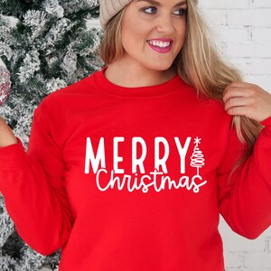 Puede incluir: Una sudadera roja con la inscripci&oacute;n blanca "MERRY Christmas" y un &aacute;rbol de Navidad estilizado. Una prenda festiva para las fiestas.