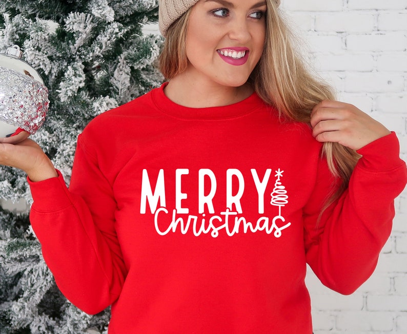 Puede incluir: Una sudadera roja con la inscripci&oacute;n blanca "MERRY Christmas" y un &aacute;rbol de Navidad estilizado. Una prenda festiva para las fiestas.