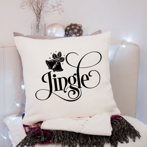 Puede incluir: Coj&iacute;n cuadrado color crema con la palabra "Jingle" en fuente negra. El coj&iacute;n presenta un dise&ntilde;o de campana y acebo negro. Debajo se ven una camisa blanca y una manta estampada, lo que sugiere un tema festivo.