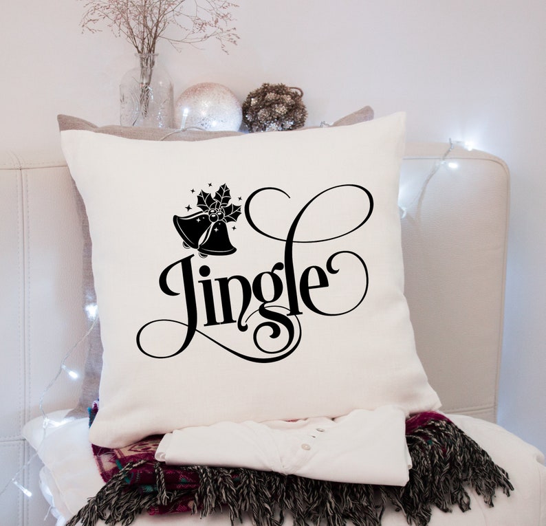 Puede incluir: Coj&iacute;n cuadrado color crema con la palabra "Jingle" en fuente negra. El coj&iacute;n presenta un dise&ntilde;o de campana y acebo negro. Debajo se ven una camisa blanca y una manta estampada, lo que sugiere un tema festivo.