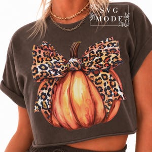 May include: A dark grey short-sleeve t-shirt featuring a graphic of an orange pumpkin adorned with a large leopard print bow. The text SVG MODE is printed in the upper right corner. The shirt has a crew neckline and a cropped hemline.